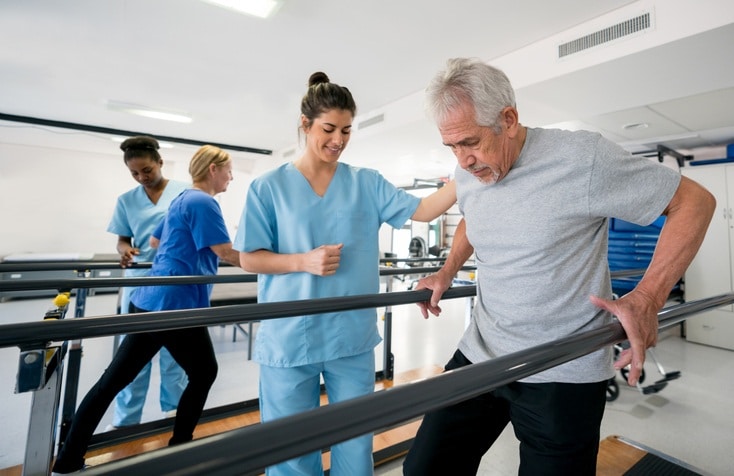 Physical therapist helping a senior walk between parallel bars